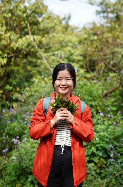 Rosie in an orange jacket holding fresh leaves on a forest trail