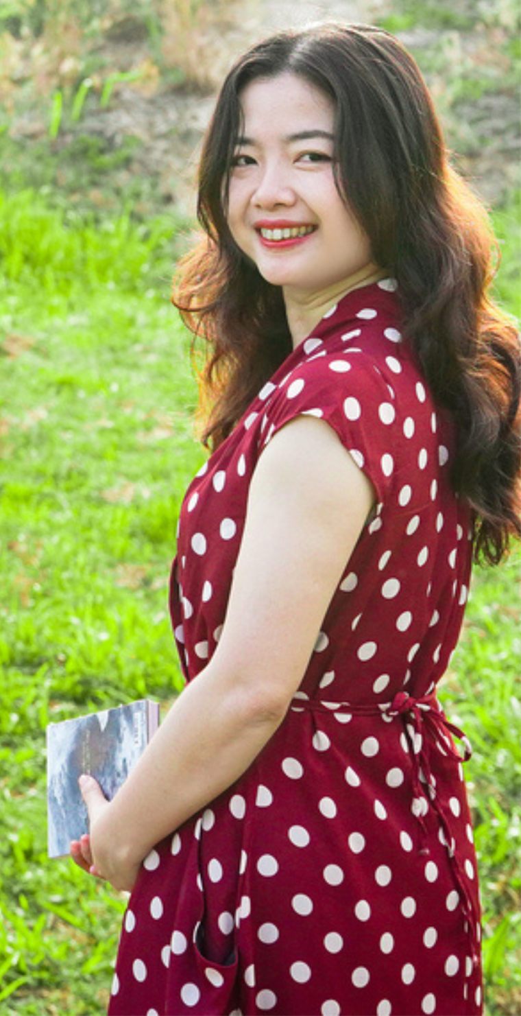 Rosie Tran smiling in a red polka-dot dress, holding a book in a green field